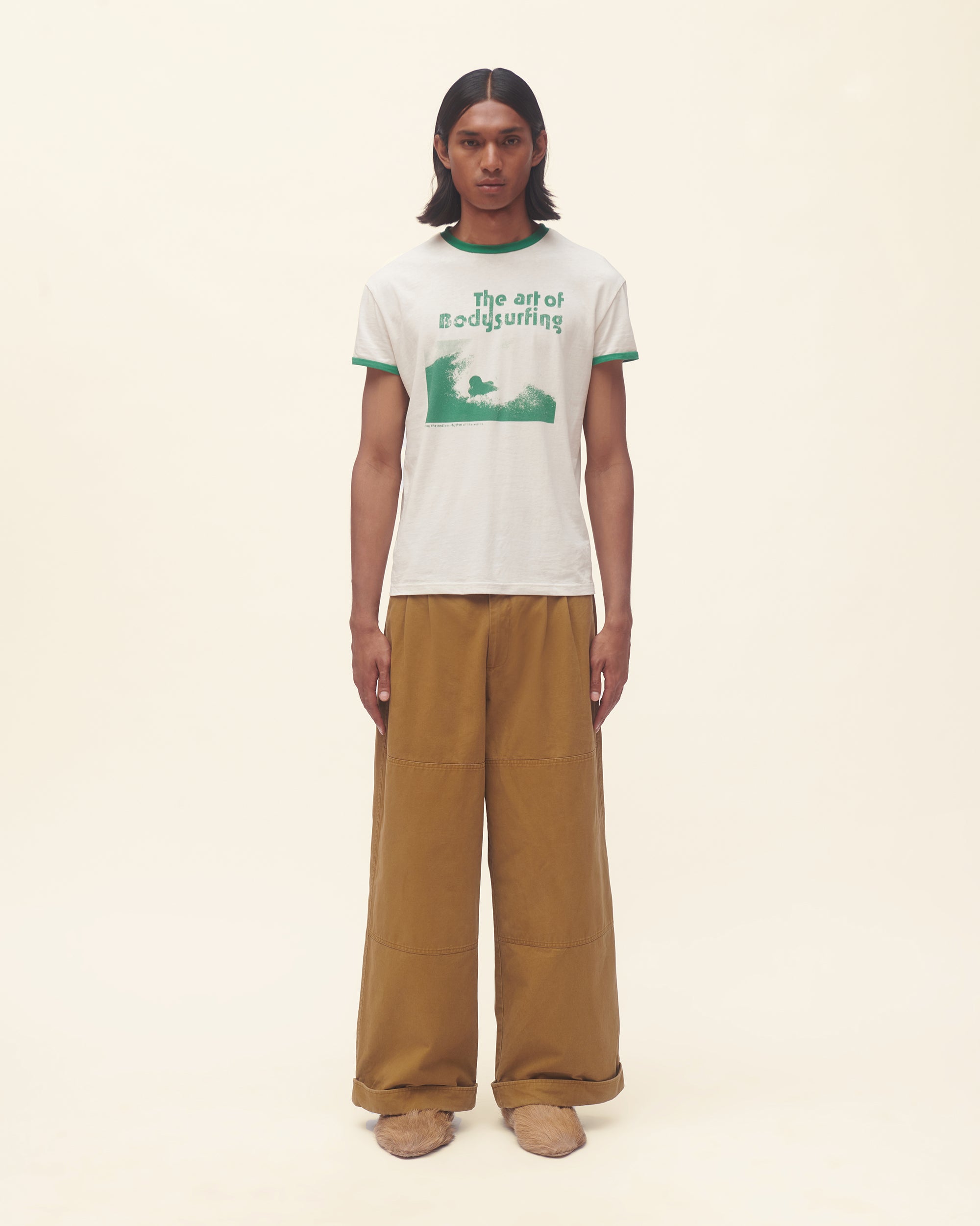 Person wearing a white t-shirt with green text and graphics, paired with brown pants on a beige background