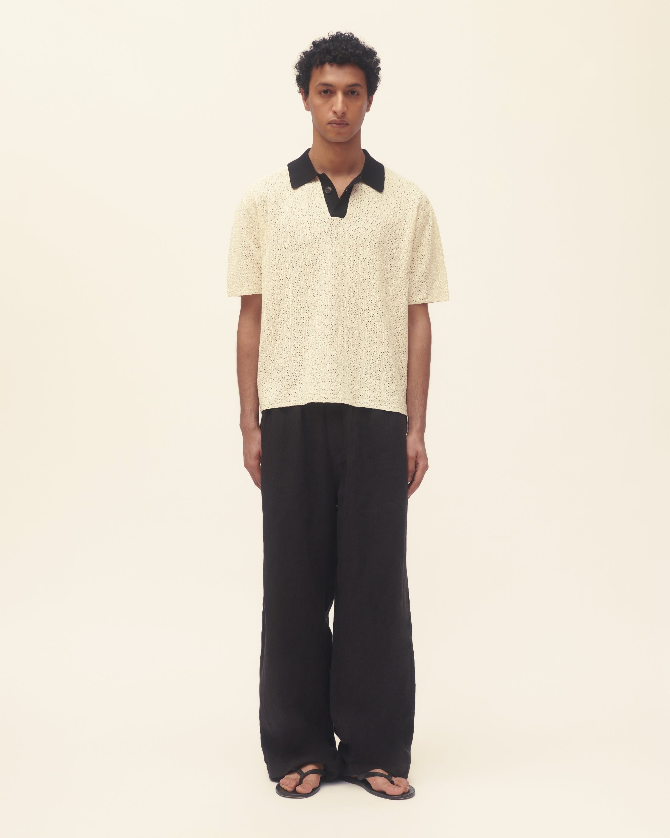 Person wearing white crochet polo with contrast black collar, styles with black linen pants on cream background.
