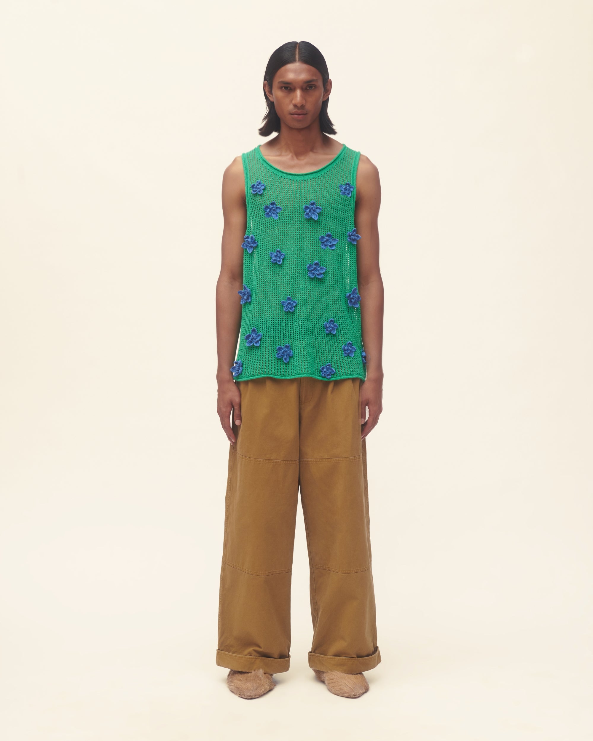 Person wearing a green sleeveless top with blue floral patterns and brown pants on a beige background