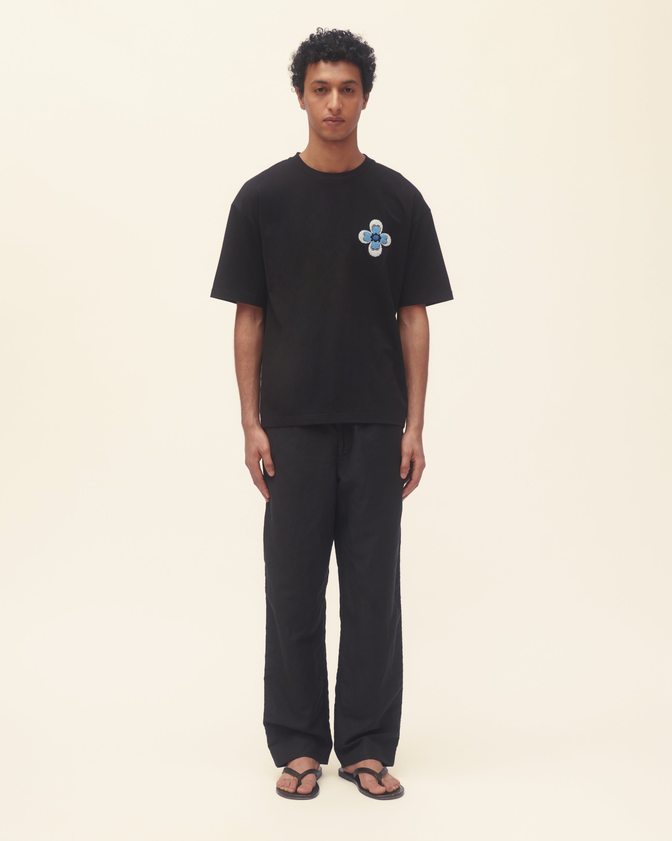 Person wearing a black t-shirt with a blue flower design and black pants on a beige background