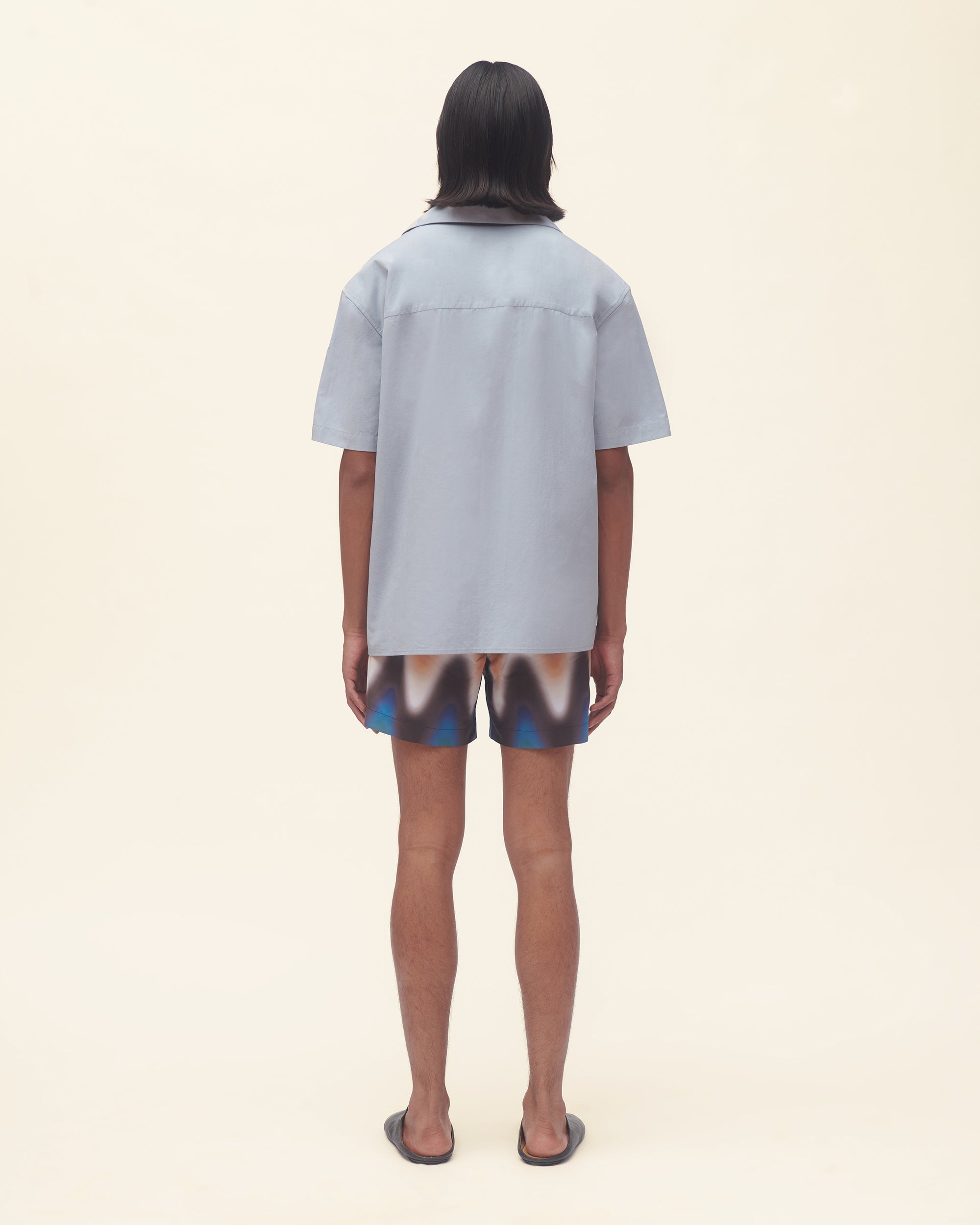Person wearing a light blue shirt and wavy print swim shorts on a beige background