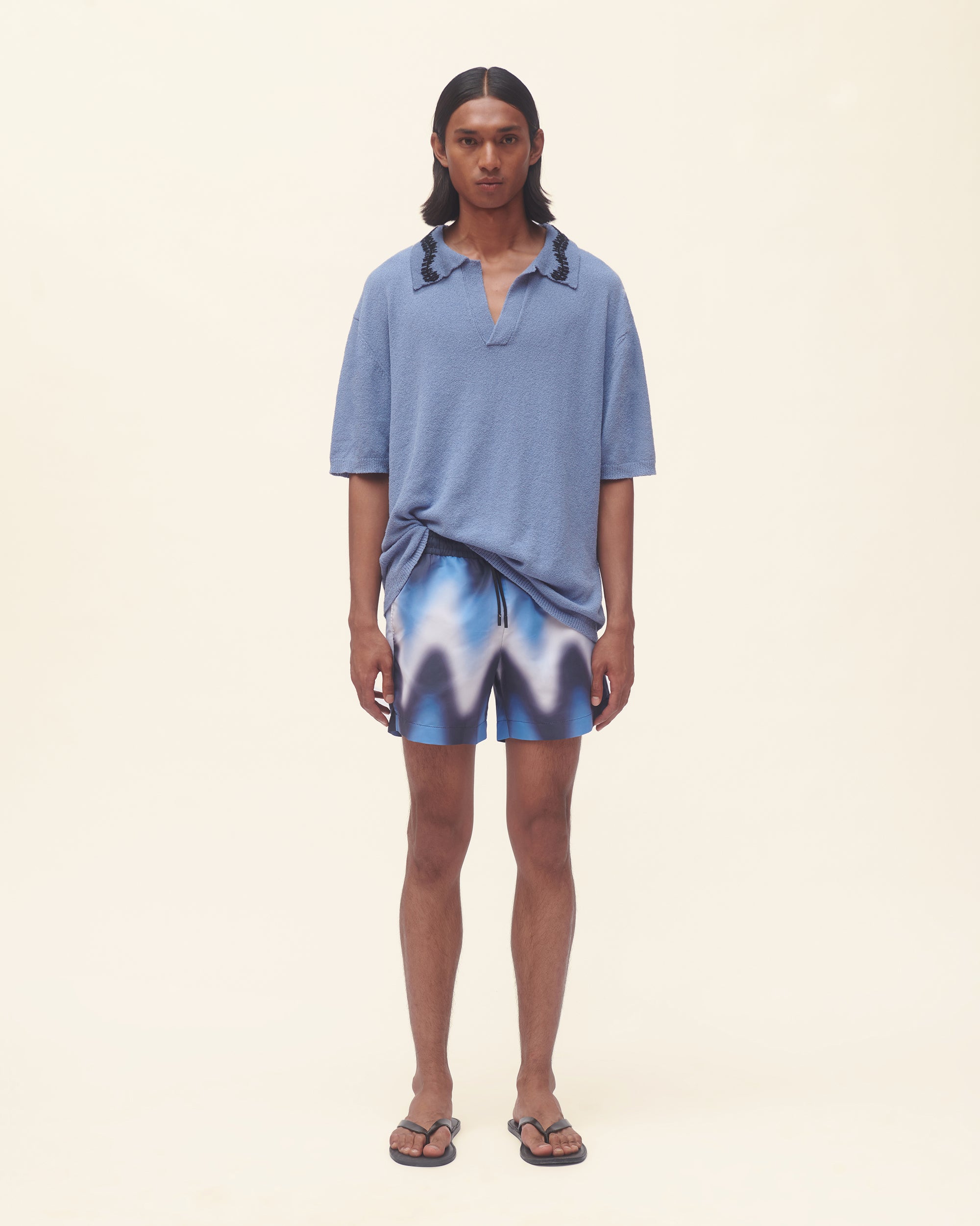Person wearing a blue polo shirt and wavy print swim shorts on a beige background
