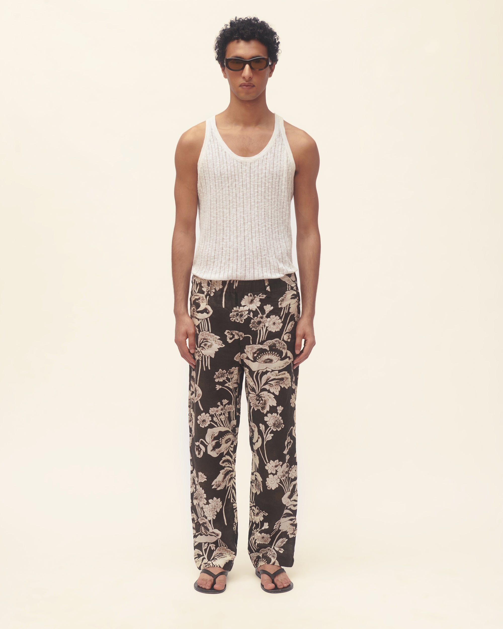 Man wearing a white tank top and black floral pants on a beige background