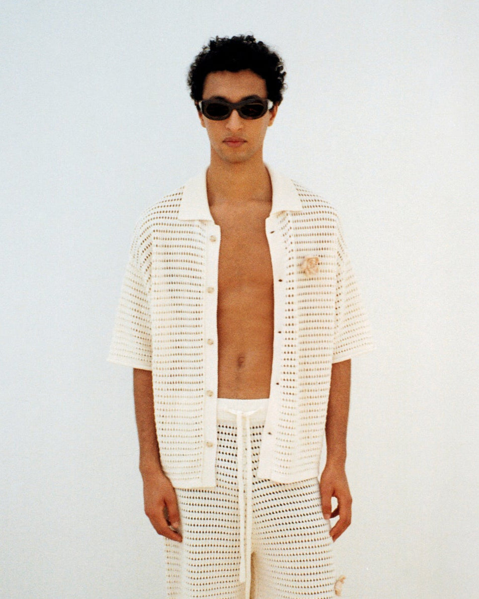 Man wearing a white knit top and shorts with sunglasses against a plain background