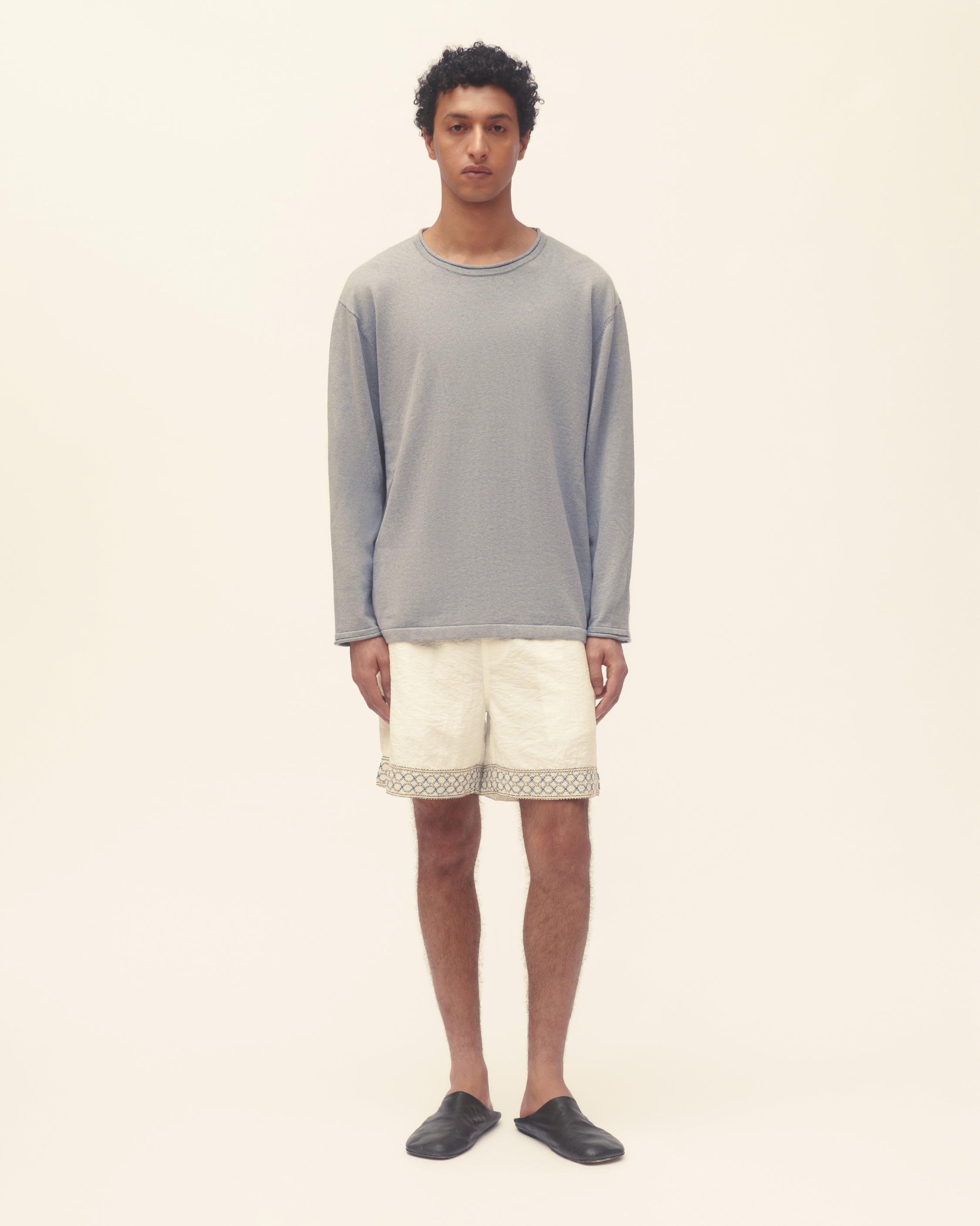 Man wearing a gray long-sleeve shirt and white shorts with lace trim on a beige background