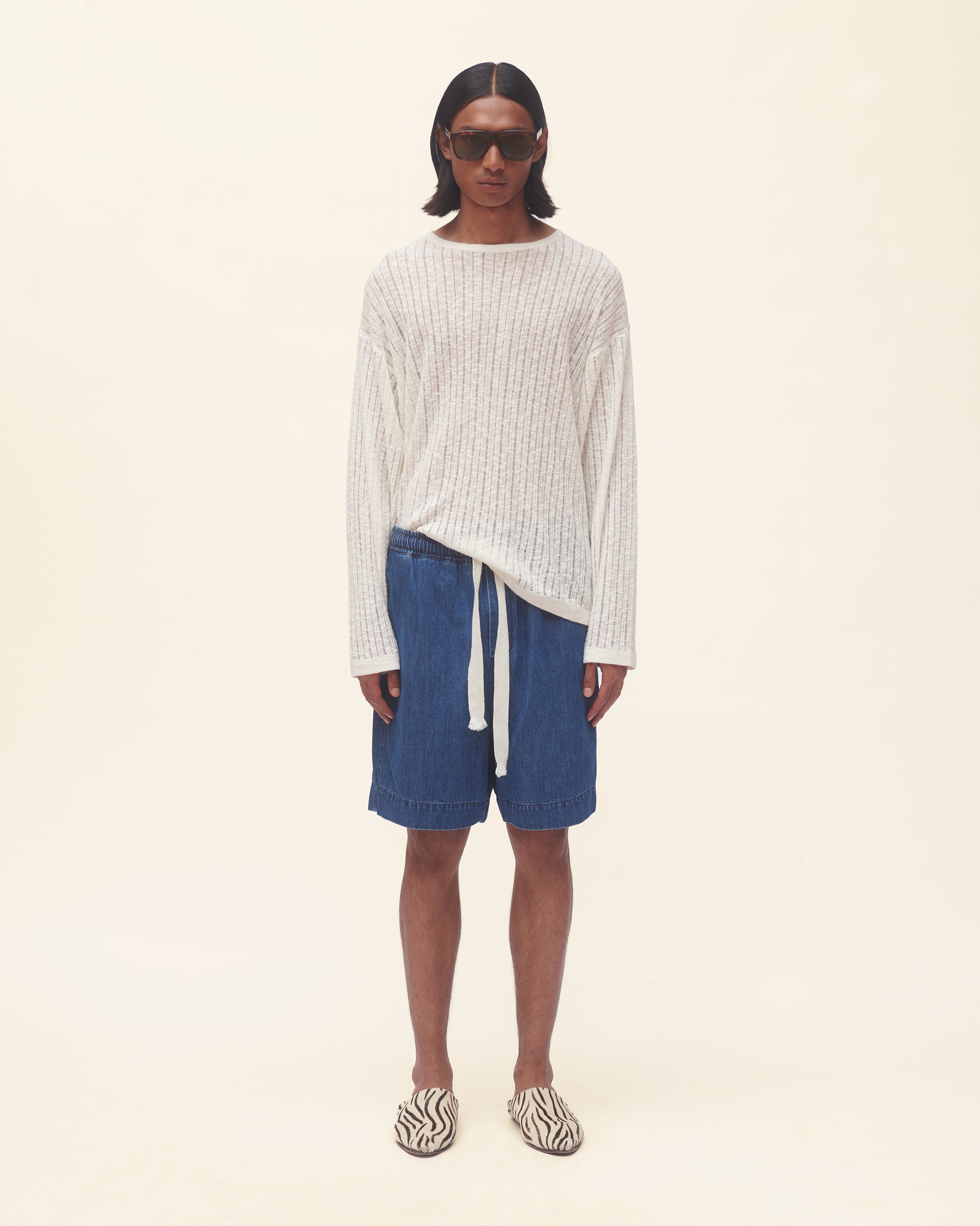 Person wearing a white ribbed sweater and blue shorts on a beige background