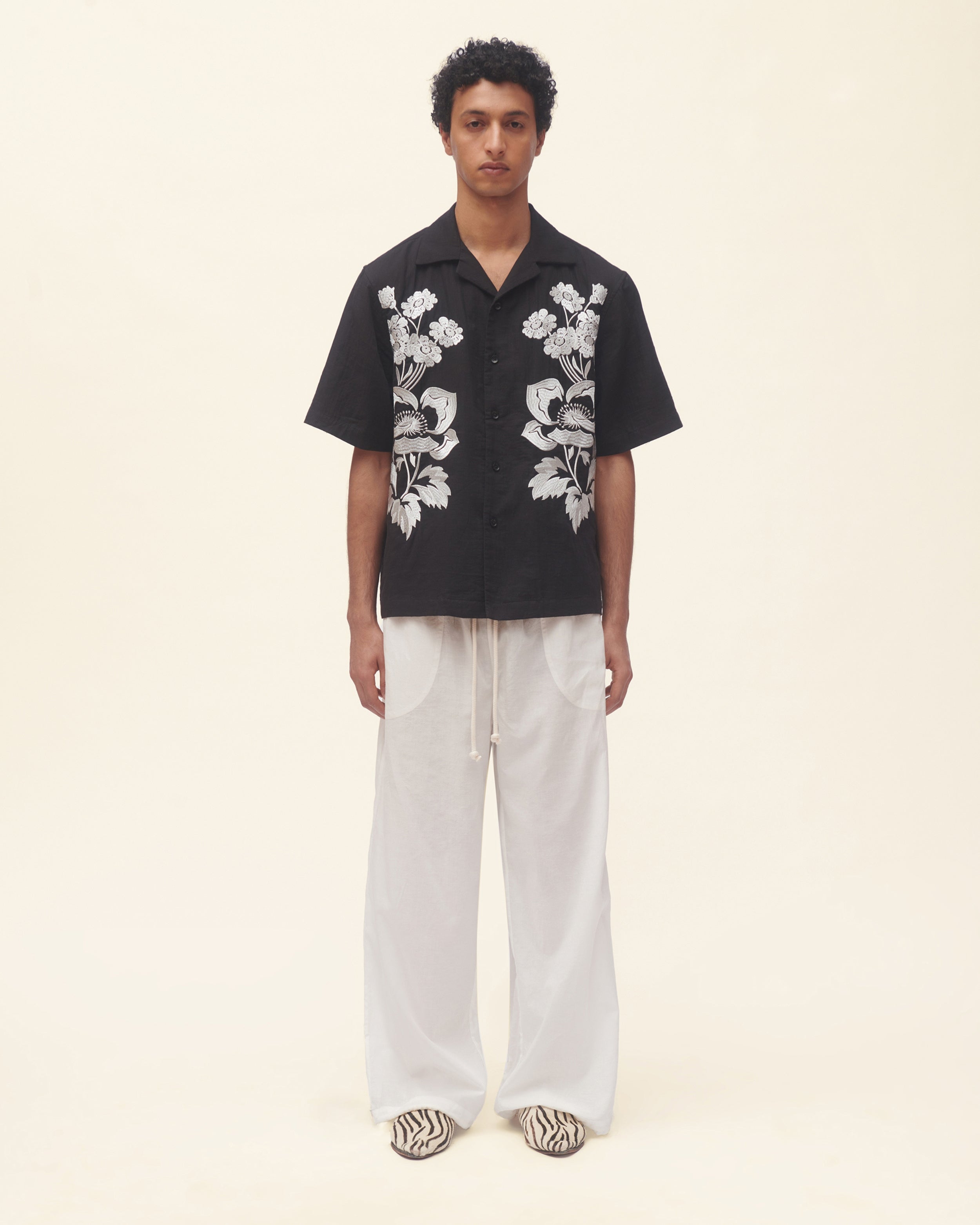 Person wearing a black shirt with white floral patterns and white pants on a beige background