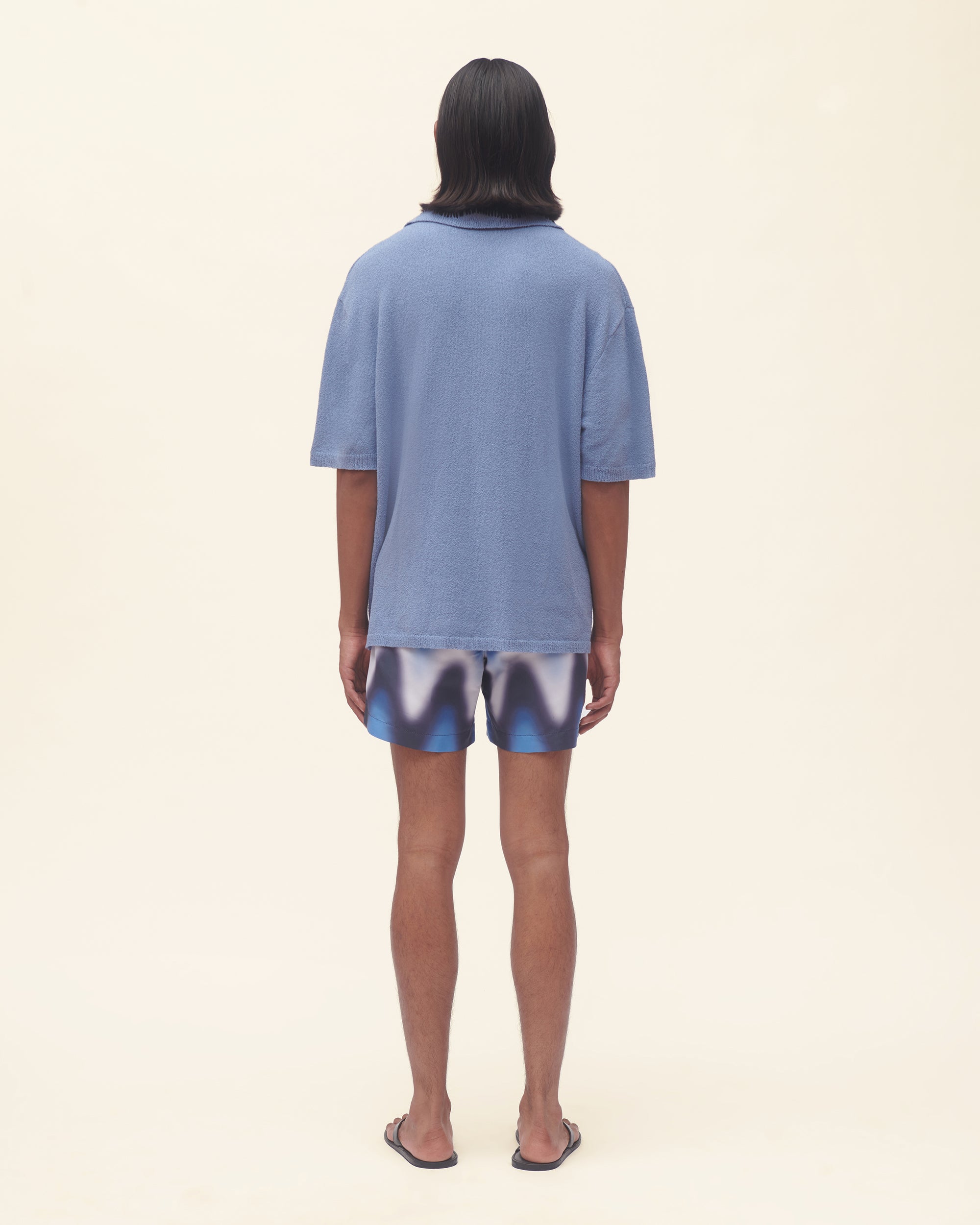 Person wearing a blue polo shirt and wavy print swim shorts on a beige background