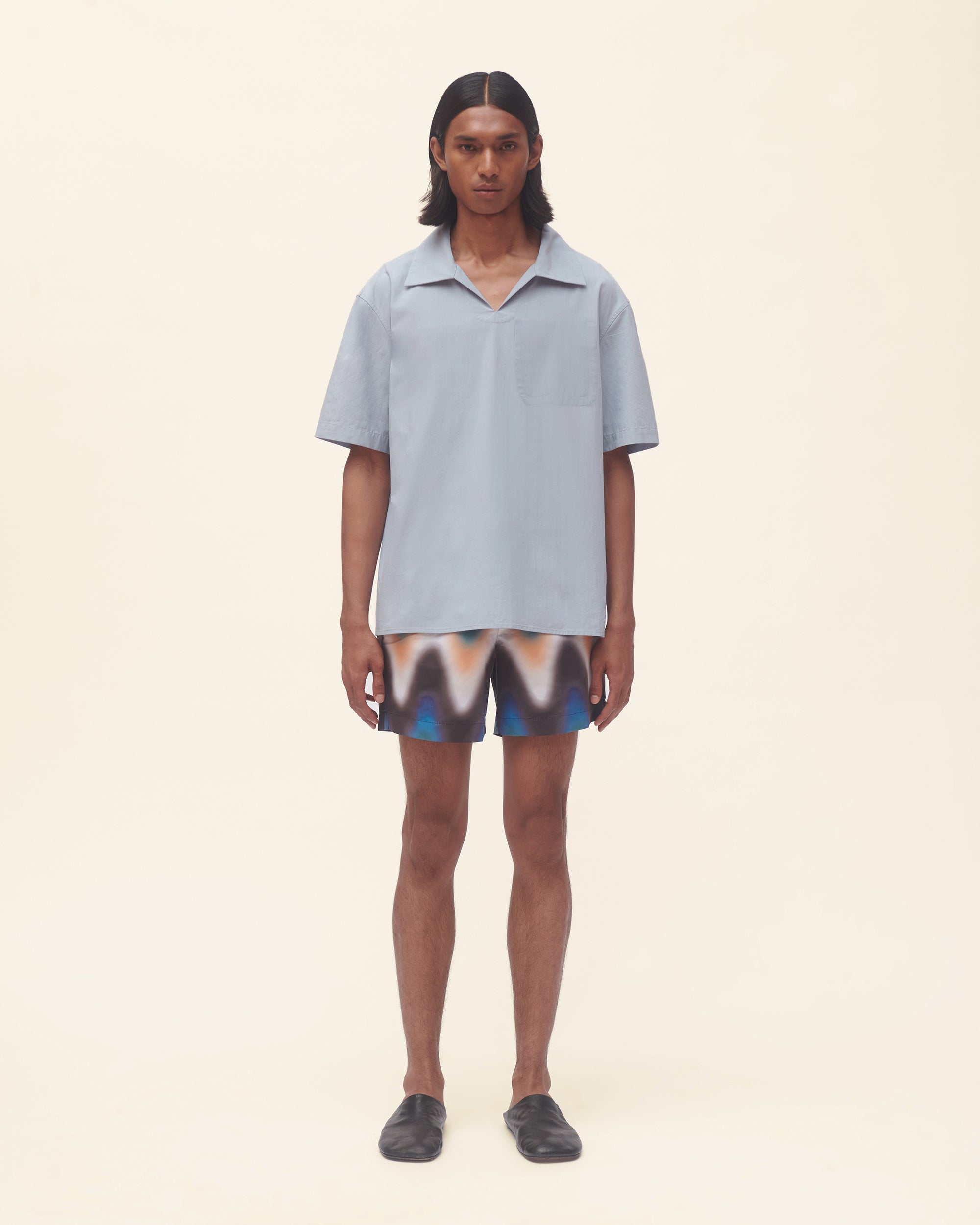 Person wearing a light blue shirt and wavy print swim shorts on a beige background