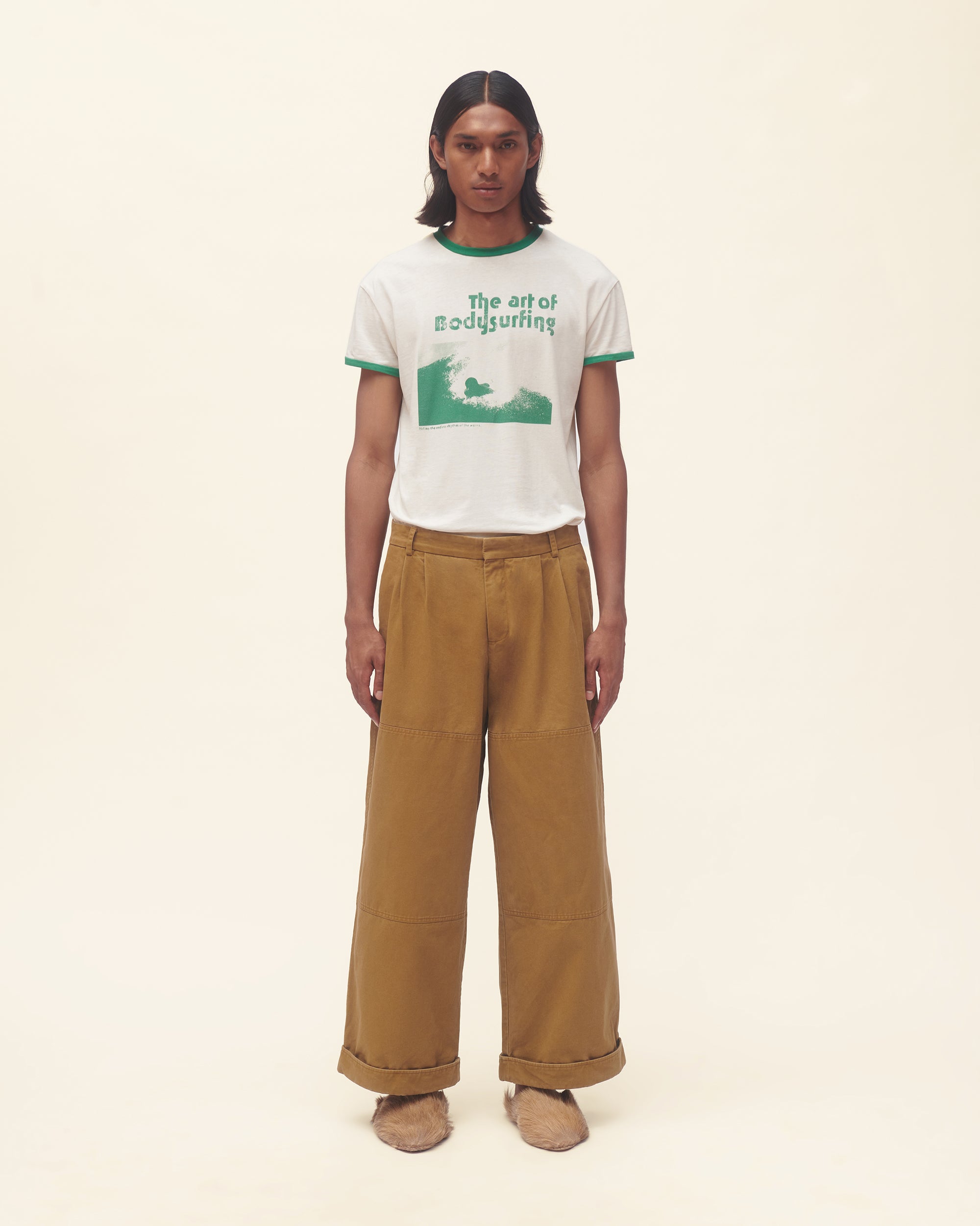 Person wearing a white t-shirt with green text and brown pants on a beige background