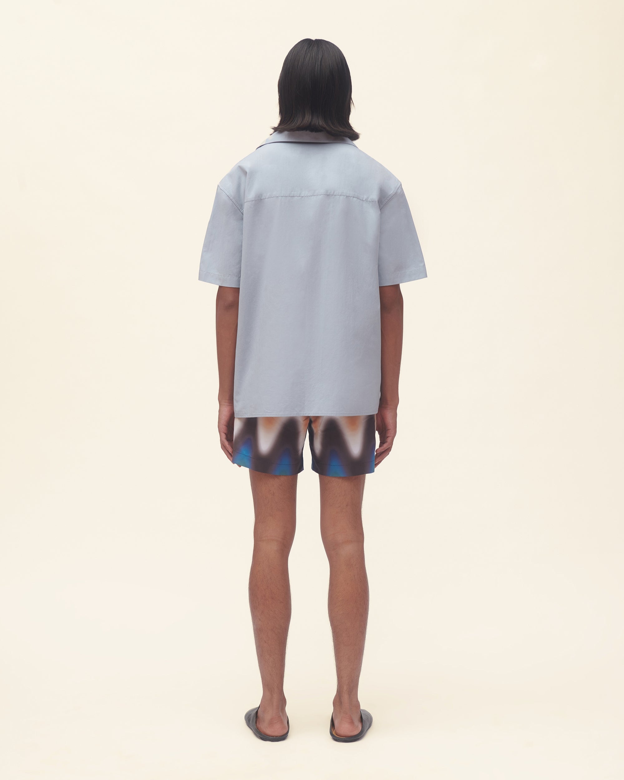 Person wearing a light blue shirt and patterned shorts on a beige background